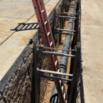 Service ladder coming out of a long ground opening with a black pipe running its length.