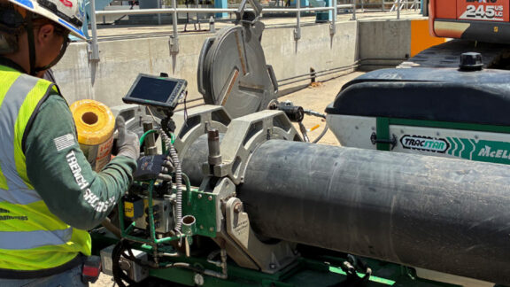 Pipeline construction worker stading next to large black pipe and referencing design plans on a tablet.