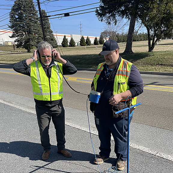 Two men in yellow worker vests and headphones testing acoustic leak detection technology.